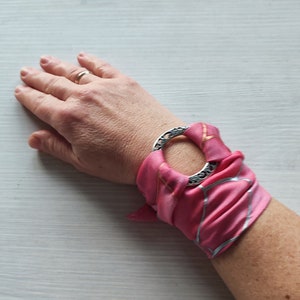 Puede incluir: Una bufanda de seda rosa con un acento de anillo plateado. La bufanda está atada alrededor de una muñeca.