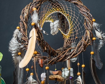 May include: A handcrafted dreamcatcher with a woven willow branch frame. The dreamcatcher features a webbed centre, adorned with feathers, wooden beads, and crystals. The colour palette includes brown, beige, grey, and yellow.