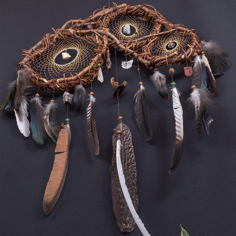 May include: A wall hanging featuring three interconnected circular frames made of twisted brown twigs. Each frame contains a black web with a central white stone. Numerous brown, grey, and white feathers hang from the frames on thin strings.