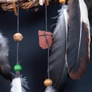 May include: A dreamcatcher featuring a woven branch hoop with hanging black strings. The strings are adorned with a textured tan bead, a smooth green bead, a polished tan bead, a flat red stone, and several long black and white feathers.
