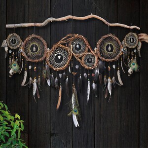 May include: A large wall hanging featuring five interconnected circular dream catchers suspended from a natural wooden branch. The hoops are wrapped in brown twigs with intricate white webbing, clear crystals, and various hanging feathers.