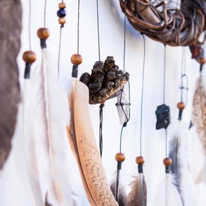 May include: A dream catcher featuring a woven branch hoop, hanging strings with wooden beads, dark brown and white feathers, a small pinecone, and a dark stone fragment. The decorative item is suspended by thin black cords against a plain background.
