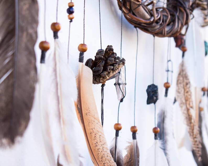 May include: A dream catcher featuring a woven branch hoop, hanging strings with wooden beads, dark brown and white feathers, a small pinecone, and a dark stone fragment. The decorative item is suspended by thin black cords against a plain background.