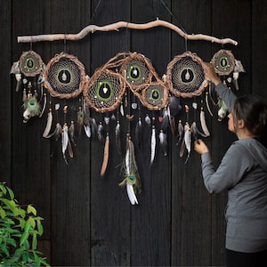 May include: A large wall hanging featuring six interconnected circular dream catchers made of brown woven branches and dark webbing. Each circle contains a central crystal, and the piece is adorned with hanging feathers and beads from a branch.