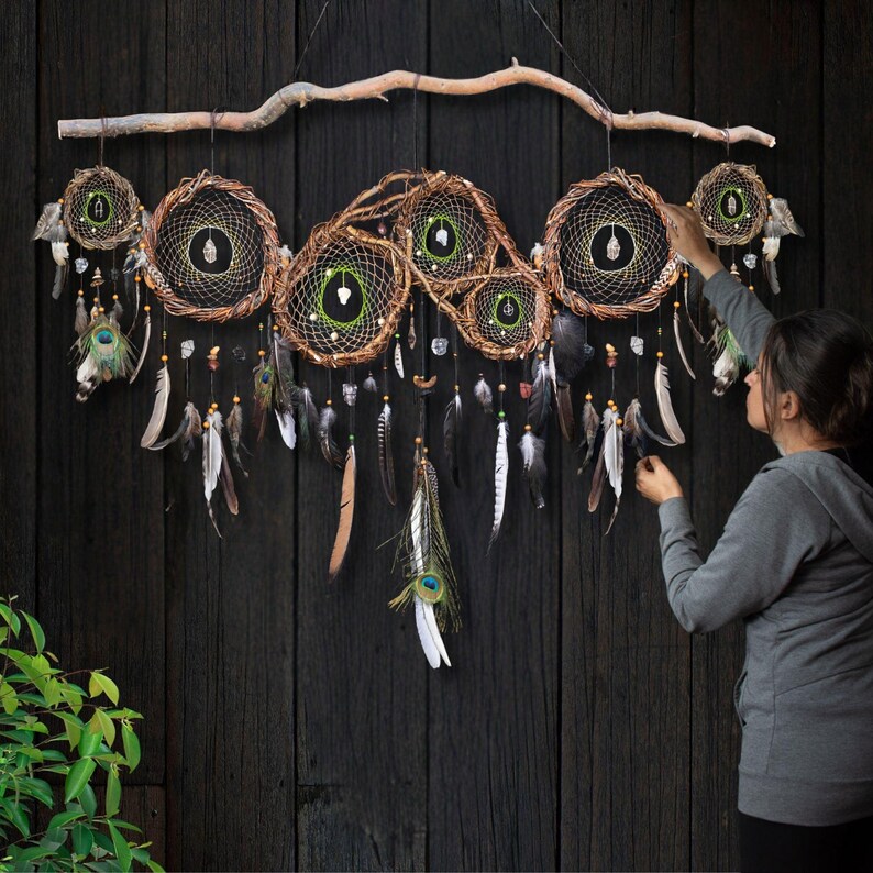 May include: A large wall hanging featuring six interconnected circular dream catchers made of brown woven branches and dark webbing. Each circle contains a central crystal, and the piece is adorned with hanging feathers and beads from a branch.