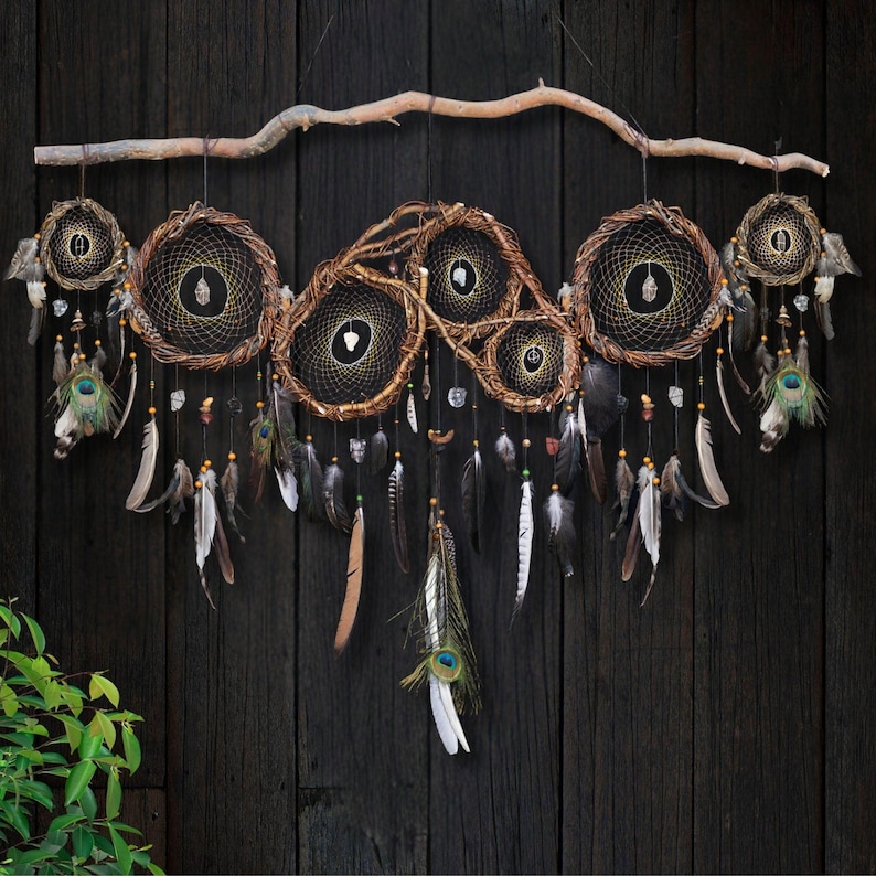 May include: A large wall hanging featuring multiple circular dream catchers woven with dark thread and brown twigs, suspended from a natural wooden branch. The piece is adorned with various hanging feathers, including peacock feathers, and small crystals.
