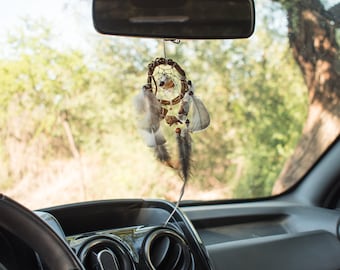 Può includere: Un acchiappasogni marrone e bianco appeso allo specchietto retrovisore di un'auto. L'acchiappasogni ha piume e perline.