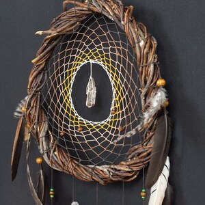 May include: A circular dreamcatcher featuring a thick, twisted brown willow branch frame. The interior web transitions from dark brown to yellow and white, centered with a clear quartz crystal. Brown wooden beads and dark feathers hang below.