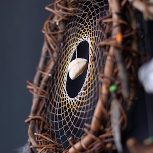 May include: A dreamcatcher featuring a circular frame made of intertwined brown twigs. The center web transitions from white to yellow, holding a single, smooth, light-colored stone suspended in the middle. Small beads and feathers are attached.