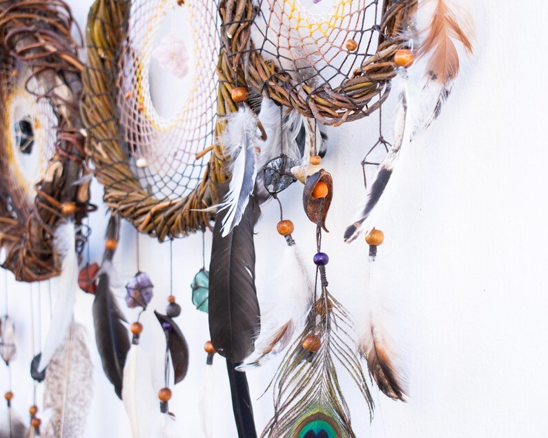 May include: A wall hanging featuring two circular dream catchers constructed from twisted brown willow branches. The webs are woven with thin thread and decorated with various natural elements including wooden beads, raw crystals, and assorted feathers.