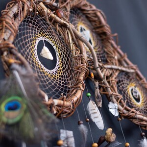 May include: A multi-hoop dreamcatcher constructed from intertwined brown twigs. The webbed centers feature a yellow and white gradient pattern with suspended white stones, small wooden beads, and feathers, including a single peacock feather.