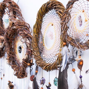 May include: A series of circular dream catchers constructed from twisted brown willow branches. Each hoop features a woven web center adorned with small crystals, wooden beads, and dangling dark feathers suspended by thin strings.
