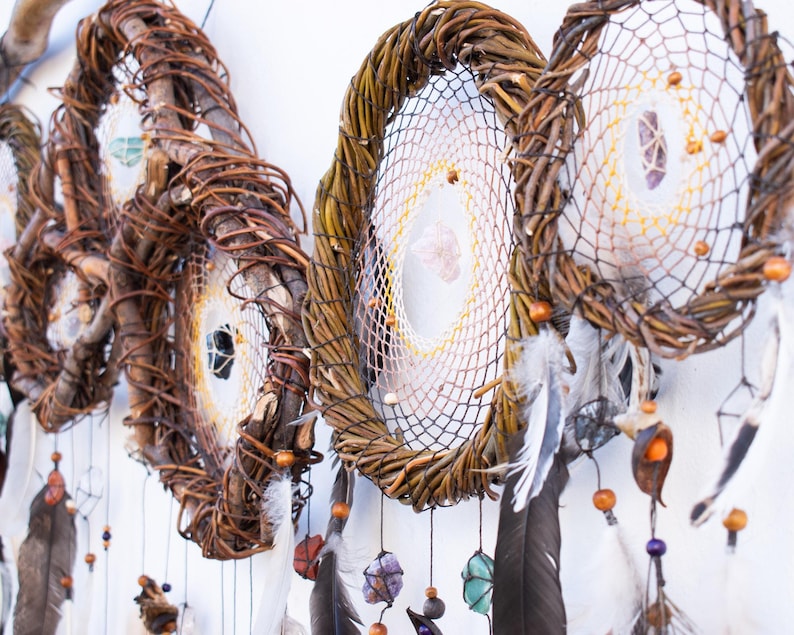 May include: A series of circular dream catchers constructed from twisted brown willow branches. Each hoop features a woven web center adorned with small crystals, wooden beads, and dangling dark feathers suspended by thin strings.