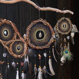 May include: A wall hanging featuring three circular woven branch hoops suspended from a horizontal wooden branch. Each hoop contains a black web with a central clear crystal, adorned with hanging feathers, beads, and a peacock eye feather.