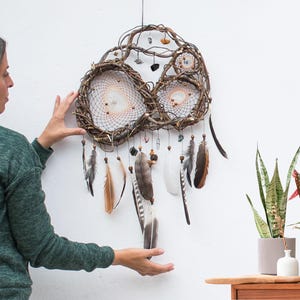 Pu&ograve; includere: Un acchiappasogni fatto a mano con una cornice in vite intrecciata, con due grandi ragnatele circolari e una pi&ugrave; piccola. &Egrave; adornato con piume nei toni del marrone, grigio e bianco, insieme a perline e piccole pietre. L'acchiappasogni &egrave; appeso a una parete bianca.
