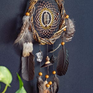 May include: A dream catcher featuring a circular woven branch frame with a web of brown and yellow thread. A clear quartz crystal hangs in the center. The piece is adorned with wooden beads, dark brown feathers, and raw crystal stones hanging from strings.