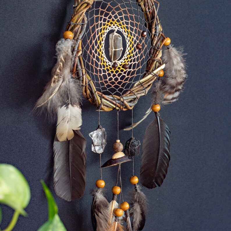 May include: A dream catcher featuring a circular woven branch frame with a web of brown and yellow thread. A clear quartz crystal hangs in the center. The piece is adorned with wooden beads, dark brown feathers, and raw crystal stones hanging from strings.