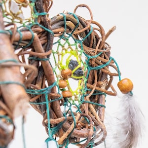 May include: Close-up of a handmade dreamcatcher. The dreamcatcher features a woven circular frame made of brown twigs, with a web of green and yellow string. Wooden beads and a grey feather are attached.