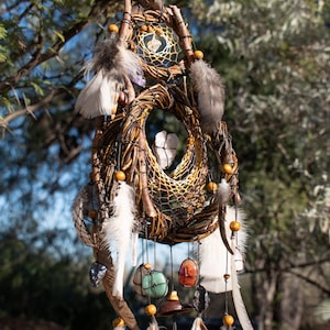 May include: A handcrafted dreamcatcher with a woven willow frame, adorned with feathers, beads, and hanging stones. The dreamcatcher features a central web design and is suspended from a tree branch, showcasing natural and earthy tones.
