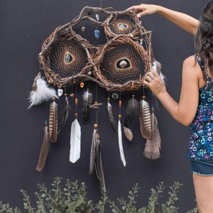 May include: A triple dream catcher with brown woven branches, white feathers, and crystals hanging from the bottom. The dream catcher is hanging on a dark grey wall.