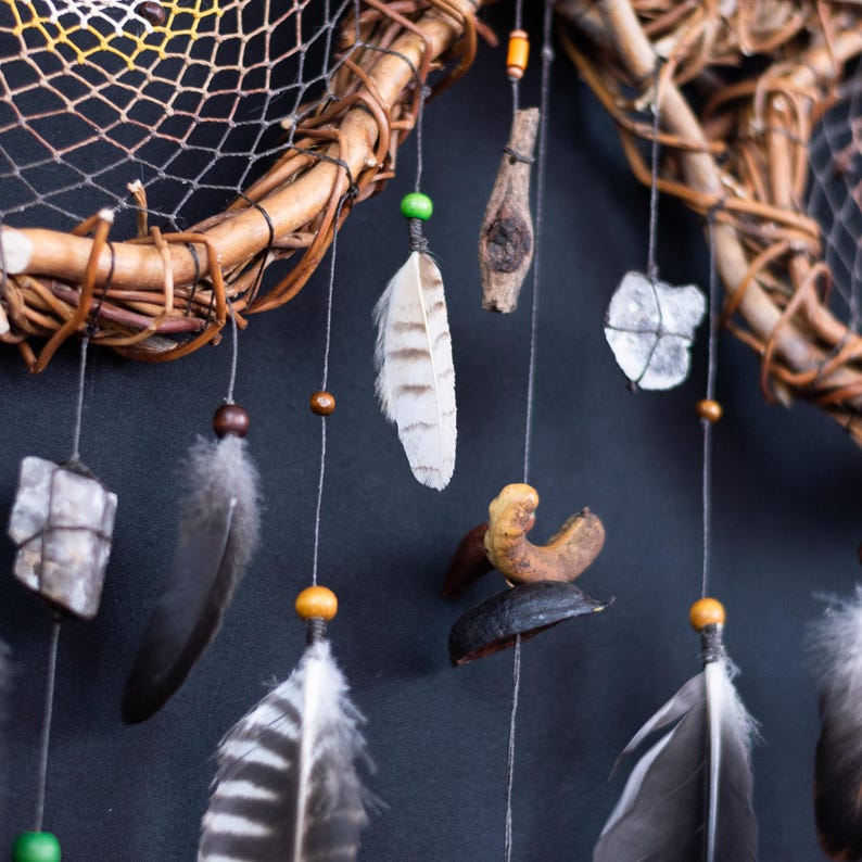 May include: A dream catcher featuring a circular woven branch frame with a web, adorned with hanging elements including grey and white striped feathers, small wooden beads, a rough grey stone, a piece of driftwood, and a dark curved seed pod.