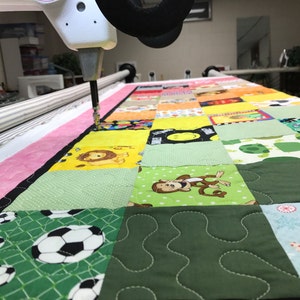 Puede incluir: Primer plano de una colcha de retazos colorida que se est&aacute; acolchando en una m&aacute;quina de acolchar de brazo largo. La colcha est&aacute; hecha de varias telas con diferentes patrones, incluyendo animales, caras sonrientes y formas geom&eacute;tricas. La m&aacute;quina est&aacute; cosiendo una tela verde con un patr&oacute;n ondulado.