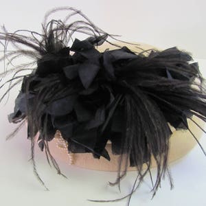 May include: A black feather hat with a large black feather trim. The hat is made of a light brown fabric.