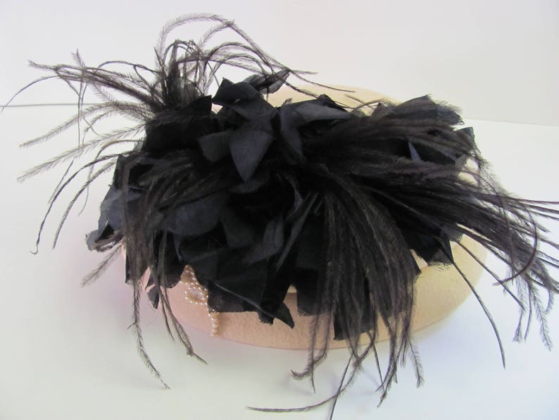 May include: A black feather hat with a large black feather trim. The hat is made of a light brown fabric.