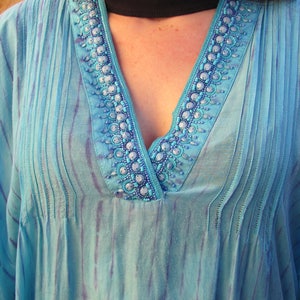 May include: A light blue, sheer tunic with a V-neckline and beaded trim. The tunic has a tie-dye pattern and is pleated in the front.