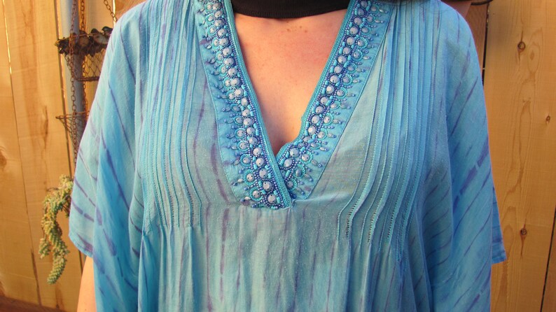 May include: A light blue, sheer tunic with a V-neckline and beaded trim. The tunic has a tie-dye pattern and is pleated in the front.