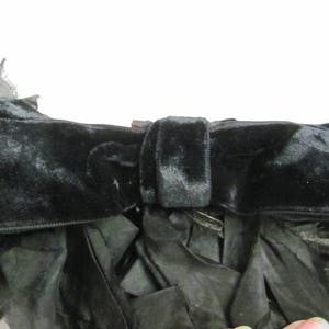 May include: A black velvet bow with a large, fluffy black feather trim. The bow is tied with a knot in the center and has a long, flowing tail.