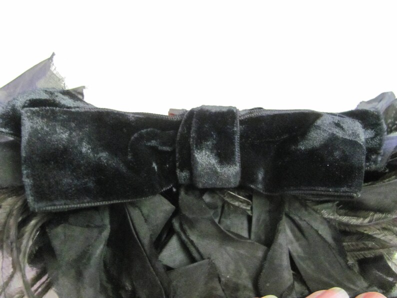 May include: A black velvet bow with a large, fluffy black feather trim. The bow is tied with a knot in the center and has a long, flowing tail.