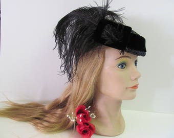 May include: A black velvet hat with a black feather and a red rose accent. The hat is on a mannequin head with long blonde hair.