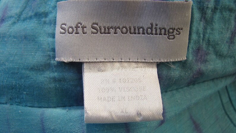 May include: A white tag with the text "Soft Surroundings" in black lettering. The tag is attached to a teal fabric with a white and blue pattern.