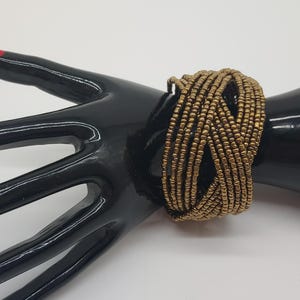 May include: A gold-colored beaded bracelet is displayed on a black hand-shaped display. The bracelet is made of many strands of small beads woven together. The display hand has a red painted fingernail.