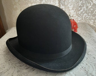 May include: A black felt bowler hat with a black grosgrain ribbon band. The hat has a rounded crown and a wide brim.