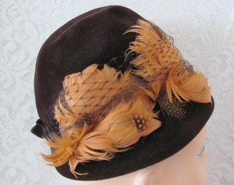 May include: A brown felt hat with a wide brim and a brown velvet band. The hat is decorated with a cluster of orange feathers and a black net.