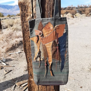 May include: A green canvas tote bag with a patchwork design featuring brown leather, burlap, and blue fabric. The bag has long straps and is hanging on a wooden post.