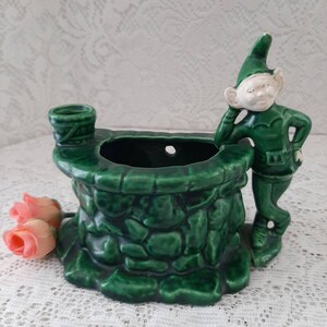 May include: Green ceramic planter shaped like a stone well with a small elf figurine leaning on the side. The planter has a hole for drainage and a small bucket on the side.