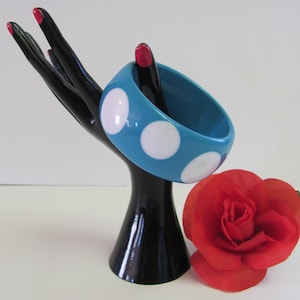 May include: A turquoise blue bangle bracelet with white polka dots. The bracelet is on a black hand-shaped display stand.