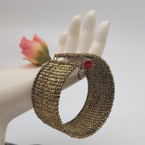 May include: A wide, gold-toned bracelet with a woven design is displayed on a white hand. A small, pink rose is placed near the bracelet. The bracelet has a small, red charm hanging from it.