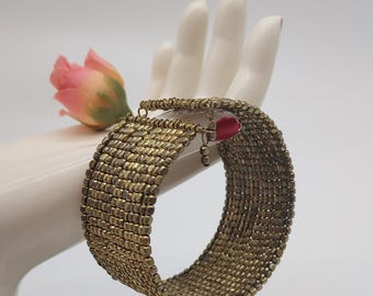 May include: A wide, gold-toned bracelet with a woven design is displayed on a white hand. A small, pink rose is placed near the bracelet. The bracelet has a small, red charm hanging from it.