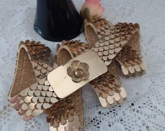 May include: A gold-tone belt with a large, ornate buckle featuring a flower design. The belt is made of woven fabric with a scaly texture.