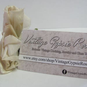May include: A white fabric rose with a business card that reads "Vintage Gypsie Rose" and "Releetle Vintage Clothing, Jewelry and Glass Ware" with the website address "www.etsy.com/shop/VintageGypsieRose" and a Facebook icon with the text "VintageGypsieRose".