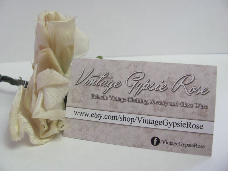 May include: A white fabric rose with a business card that reads "Vintage Gypsie Rose" and "Releetle Vintage Clothing, Jewelry and Glass Ware" with the website address "www.etsy.com/shop/VintageGypsieRose" and a Facebook icon with the text "VintageGypsieRose".