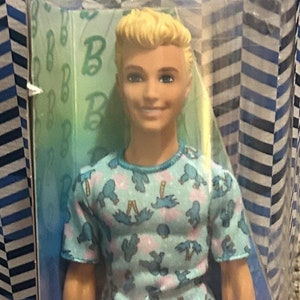 May include: A Barbie doll with blonde hair wearing a blue and white patterned shirt and white trousers. The doll is packaged in a clear plastic box with a pink Barbie logo. The box also has a warning label that says "Warning: Choking Hazard - Small parts. Not for children under 3 years."