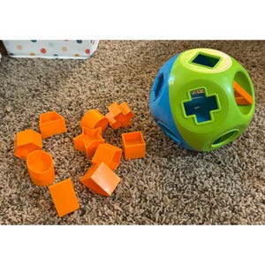 May include: A blue and green shape sorter ball with various shaped holes. Orange shapes, including a star, cube, and cylinder, are scattered around the ball. The toy is designed for early childhood development.