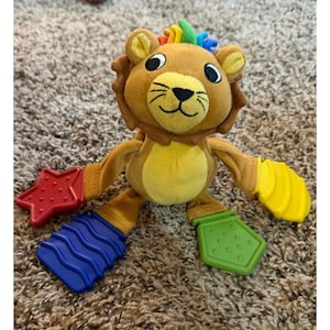 Può includere: Un giocattolo di peluche a forma di leone con criniera marrone, pancia gialla e accenti colorati. Il leone ha un ciuffo di capelli arcobaleno e appendici multicolori, tra cui una stella rossa, una forma ondulata blu e forme gialle e verdi.