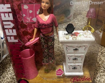 May include: Barbie Dress Up Dresser playset with a pink and white dresser, a pink trash can, and a doll wearing a pink shirt and floral pants. The box has the text "Dress Up Dresser" and "Barbie" on it.
