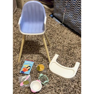 May include: A doll's high chair with a pale blue seat and wooden legs. Accessories include a white tray, a bowl, a spoon, a bottle, a bib, and a book titled "Babysitting".